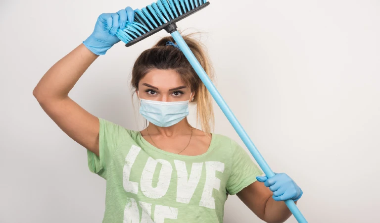 Young housewife with facemask holding tip of broom on white wall.
