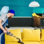 Young man with vacuum cleaner washing with vapor yellow sofa and woman wipes cuisine furniture