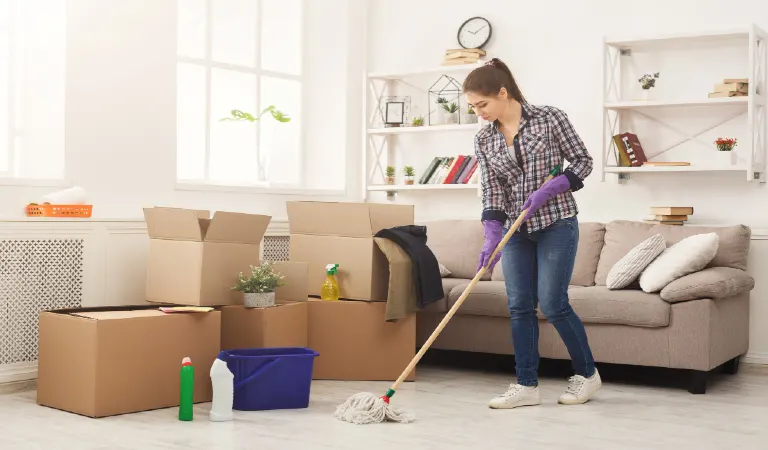 Girl with various detergents, rags and mops tiding up in living room full of carton boxes after moving to new appartment