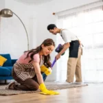 Young man and woman cleaning house indoors together happy couple vacuuming messy dirty floor for housekeeping