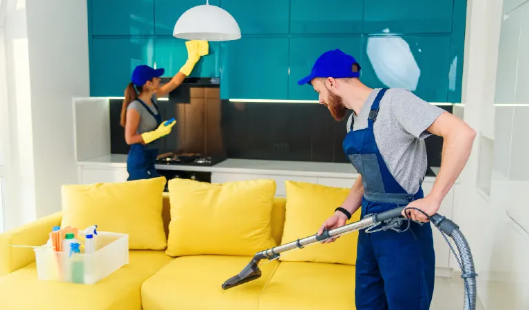 Young man vacuums yellow sofa and a girl wiping kitchen furniture. Professional cleaners in apartment