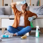 Tired woman cleaning and sitting on living room floor with detergent chemicals or sanitizer spray for hygiene at home